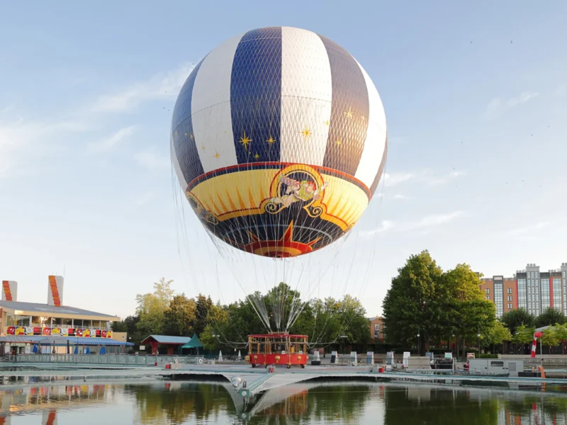 Une montgolfière captive décorée d'un motif de cirque plane au-dessus d'un étang près d'arbres, de bâtiments et d'un petit parc d'attractions à Disney Village, offrant une expérience magique à tous les visiteurs.