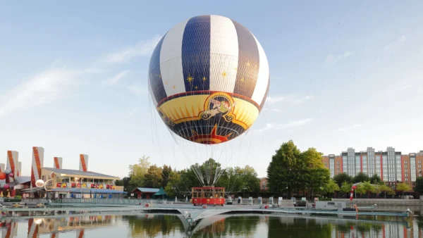 Une montgolfière captive décorée d'un motif de cirque plane au-dessus d'un étang près d'arbres, de bâtiments et d'un petit parc d'attractions à Disney Village, offrant une expérience magique à tous les visiteurs.