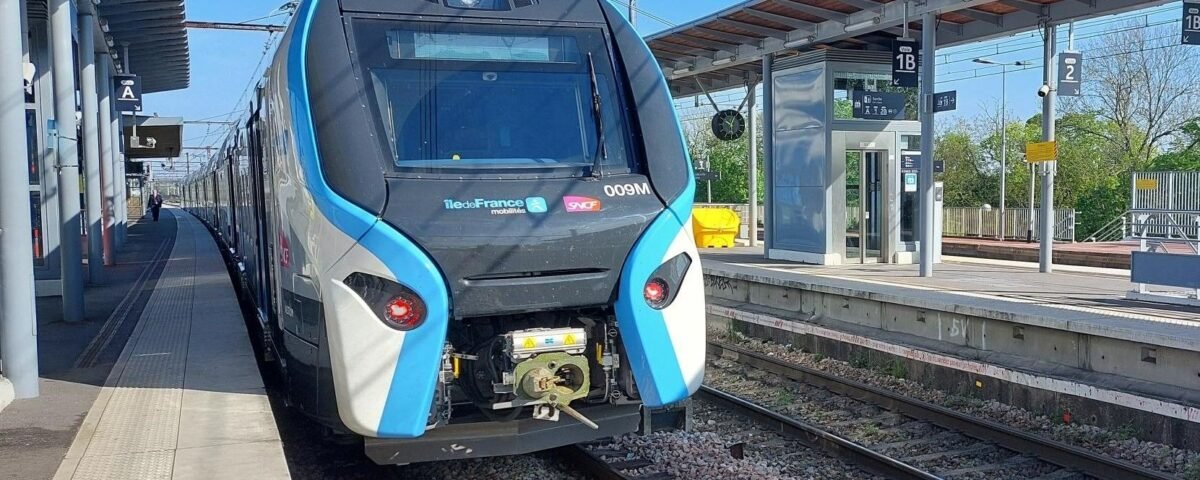 Un train de banlieue moderne est arrêté sur un quai de la gare de Marne-la-Vallée Chessy. Le train a un design épuré aux couleurs bleu et blanc. La gare, partiellement couverte par un toit, est équipée de plusieurs panneaux pour faciliter la navigation vers Val d'Europe et d'autres destinations.