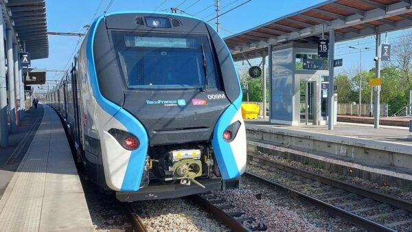 Un train de banlieue moderne est arrêté sur un quai de la gare de Marne-la-Vallée Chessy. Le train a un design épuré aux couleurs bleu et blanc. La gare, partiellement couverte par un toit, est équipée de plusieurs panneaux pour faciliter la navigation vers Val d'Europe et d'autres destinations.