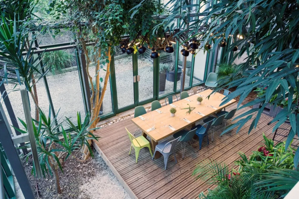 Une longue table à manger en bois avec des chaises est installée dans une serre luxuriante avec de grandes fenêtres et une verdure abondante, rappelant l'ambiance sereine que l'on retrouve dans les endroits gastronomiques autour des restaurants Val d'Europe.