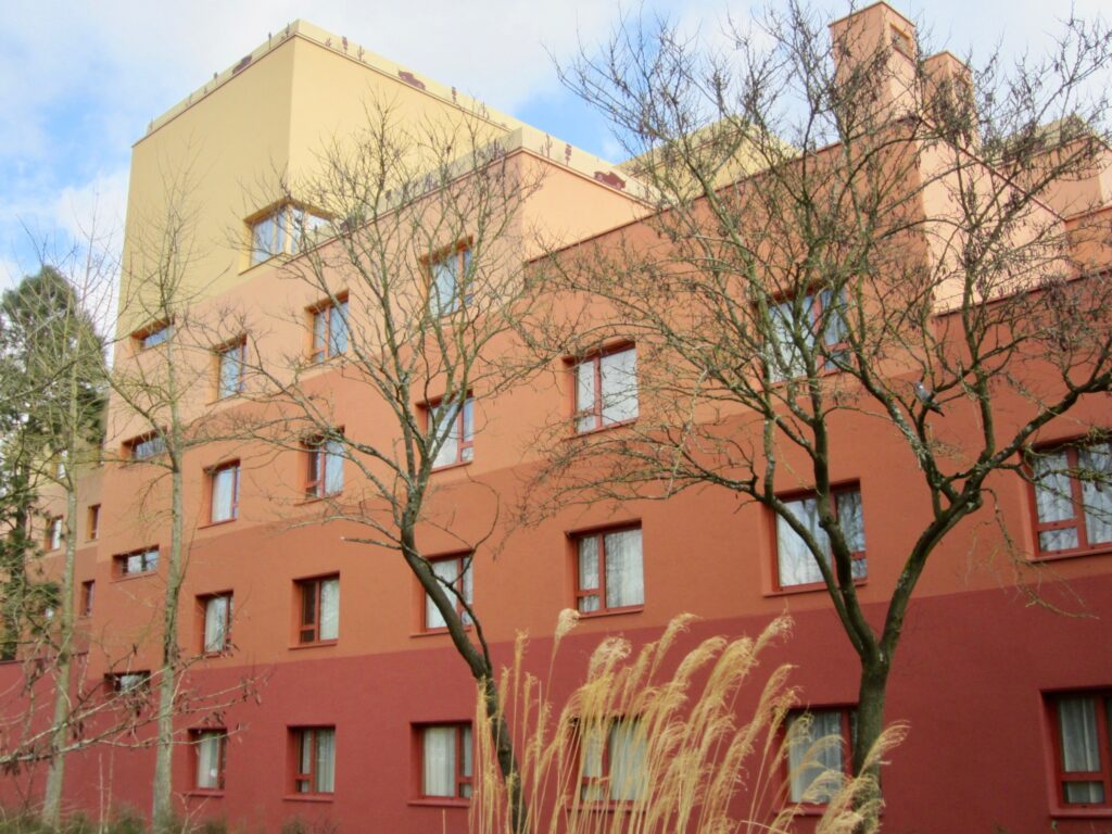 Un bâtiment à plusieurs étages aux murs rouge pâle et couleur pêche dans un quartier résidentiel, rappelant le design charmant du Disney Hotel Santa Fe, flanqué d'arbres nus et de hautes herbes au premier plan.