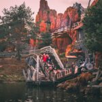 Les gens montent à bord d'un train de montagnes russes Big Thunder Mountain à travers un paysage rocheux et montagneux avec des arbres et de l'eau au premier plan.