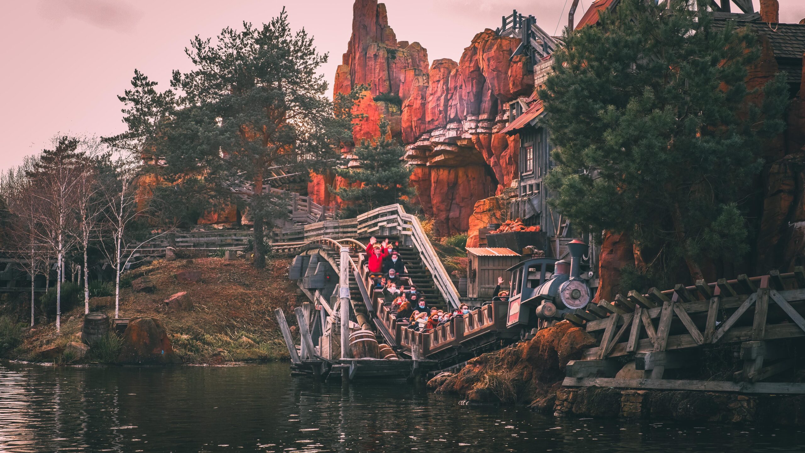 Les gens montent à bord d'un train de montagnes russes Big Thunder Mountain à travers un paysage rocheux et montagneux avec des arbres et de l'eau au premier plan.