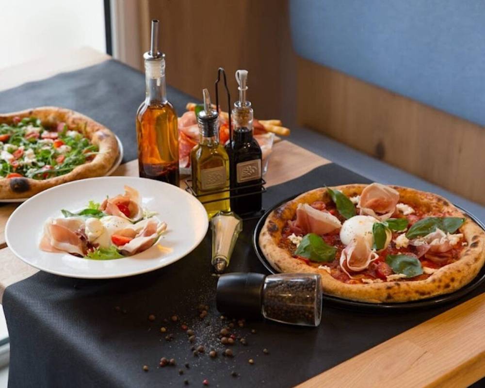 Deux pizzas et une salade sur une table du restaurant Goma, accompagnées de plusieurs bouteilles d'huile et de vinaigre, d'un moulin à poivre et de grains de poivre éparpillés. L'arrière-plan comprend une partie d'un banc bleu rembourré, reflétant l'ambiance chaleureuse caractéristique des restaurants de Val d'Europe.