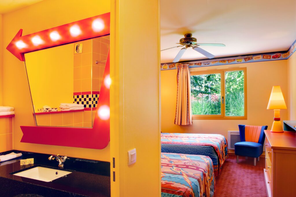 Chambre d'hôtel aux couleurs vives avec lits jumeaux, grand miroir de maquillage avec lumières, ventilateur de plafond et fenêtre donnant sur la verdure. La chambre du Disney Hotel Santa Fe présente une décoration jaune et rouge vif avec une literie à motifs. Démarrez votre moteur pour un séjour inoubliable !