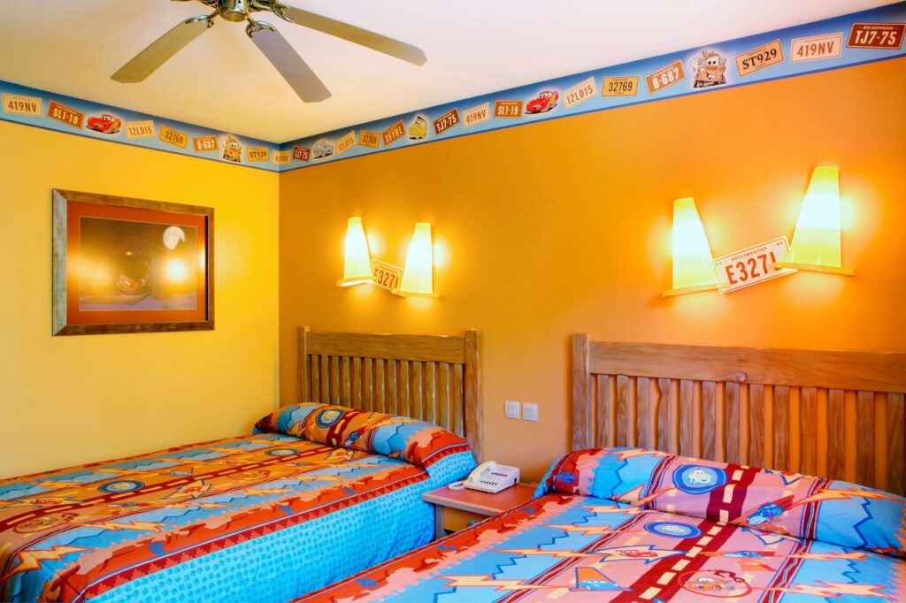 Une chambre aux couleurs vives avec deux lits en bois et une literie sur le thème des voitures vous attend à l'hôtel Santa Fe. La chambre est dotée de murs jaunes et orange avec des bordures de plaques d'immatriculation de voitures, d'un ventilateur de plafond et de lumières murales au-dessus de chaque lit. Démarrez votre moteur pour un séjour inoubliable !