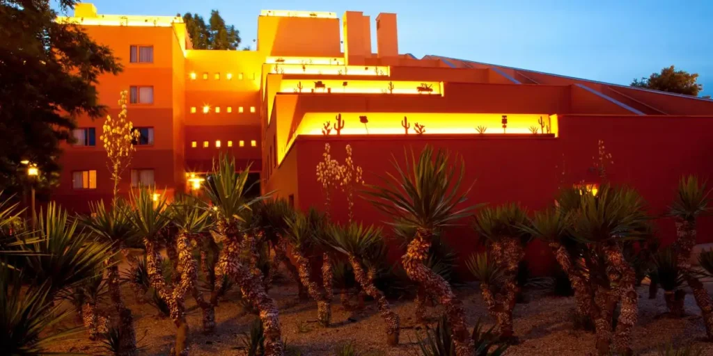 Bâtiment rouge et jaune aux fenêtres éclairées au crépuscule, entouré de plantes du désert au premier plan, rappelant l'hôtel Santa Fe.