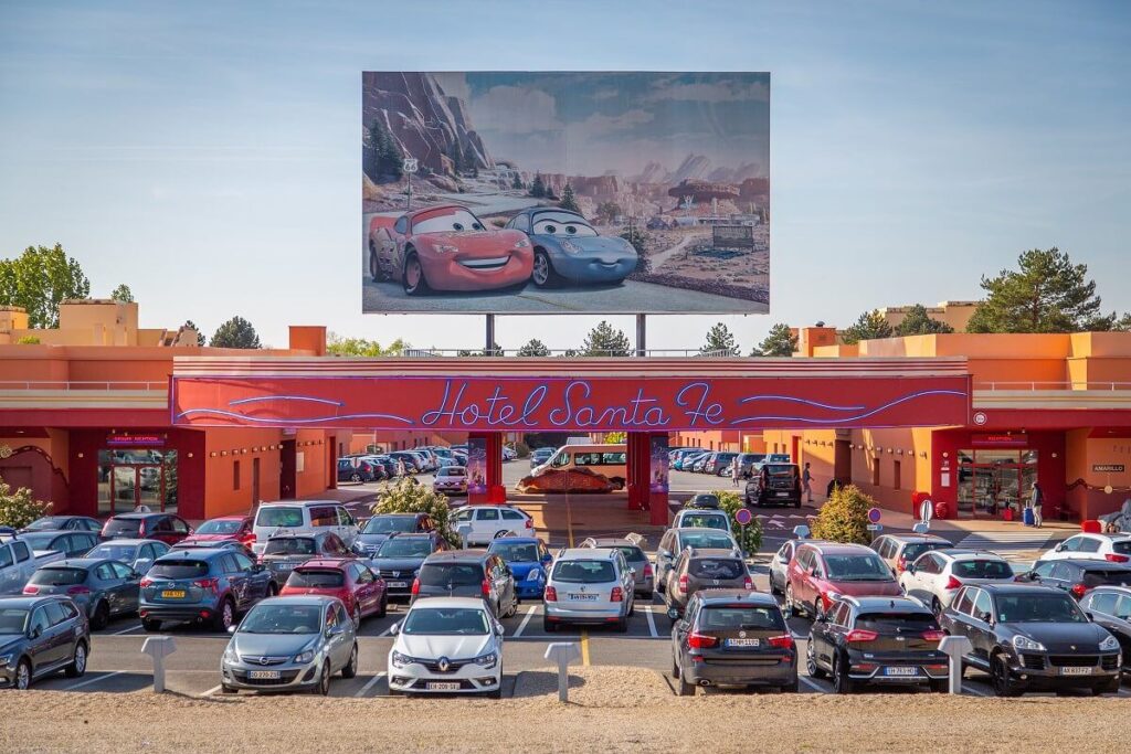 Un parking rempli de voitures devant le Disney Hotel Santa Fe, avec en arrière-plan un grand panneau publicitaire représentant les personnages du film "Cars". Démarrez votre moteur et faites de votre séjour un moment inoubliable !