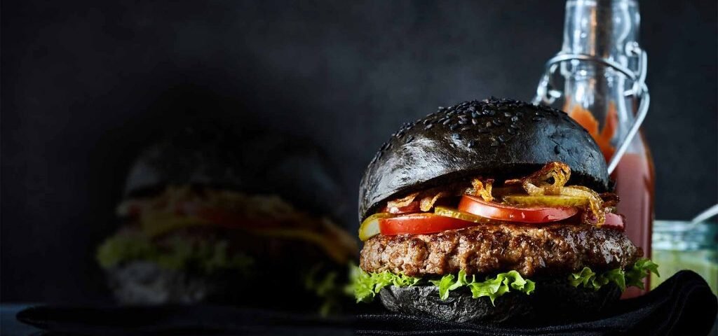 Un burger au pain noir et foncé, garni d'une galette de bœuf, de laitue, de tomates et d'oignons croustillants, est placé devant une bouteille de sauce et un pot, sur un fond sombre. Ce délice gourmand capture l'essence d'une expérience culinaire inoubliable dans les restaurants Val d'Europe.