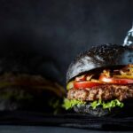 Un burger au pain noir et foncé, garni d'une galette de bœuf, de laitue, de tomates et d'oignons croustillants, est placé devant une bouteille de sauce et un pot, sur un fond sombre. Ce délice gourmand capture l'essence d'une expérience culinaire inoubliable dans les restaurants Val d'Europe.