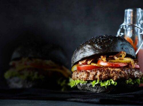 Un burger au pain noir et foncé, garni d'une galette de bœuf, de laitue, de tomates et d'oignons croustillants, est placé devant une bouteille de sauce et un pot, sur un fond sombre. Ce délice gourmand capture l'essence d'une expérience culinaire inoubliable dans les restaurants Val d'Europe.