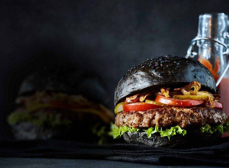 Un burger au pain noir et foncé, garni d'une galette de bœuf, de laitue, de tomates et d'oignons croustillants, est placé devant une bouteille de sauce et un pot, sur un fond sombre. Ce délice gourmand capture l'essence d'une expérience culinaire inoubliable dans les restaurants Val d'Europe.