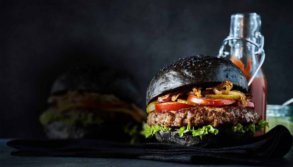 Un burger au pain noir et foncé, garni d'une galette de bœuf, de laitue, de tomates et d'oignons croustillants, est placé devant une bouteille de sauce et un pot, sur un fond sombre. Ce délice gourmand capture l'essence d'une expérience culinaire inoubliable dans les restaurants Val d'Europe.