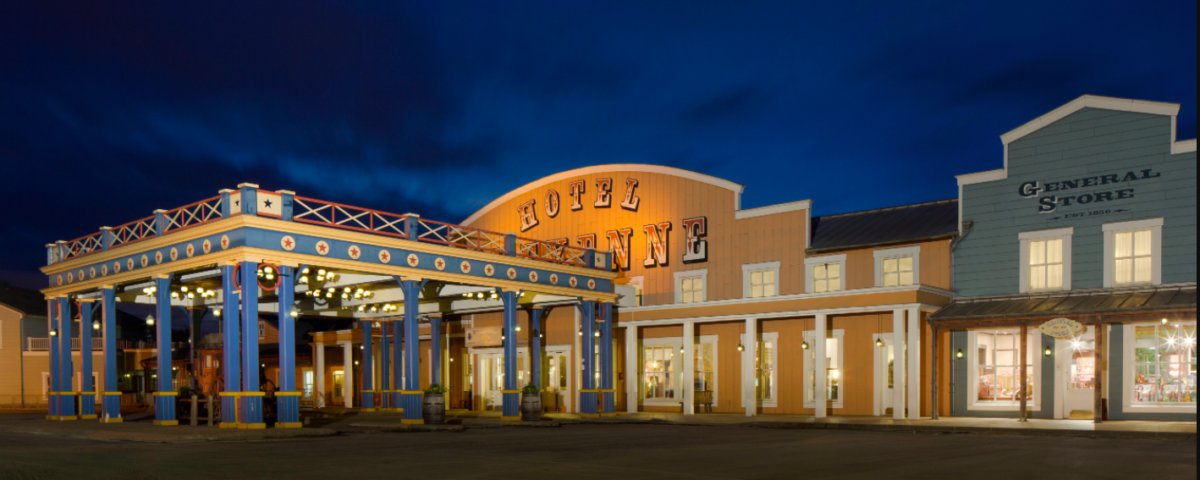 Vue nocturne d'une place de ville à thème western, avec un bâtiment étiqueté « Hôtel Cheyenne Disney » et un « Magasin général », avec des guirlandes lumineuses illuminant la zone, promettant un séjour mémorable.