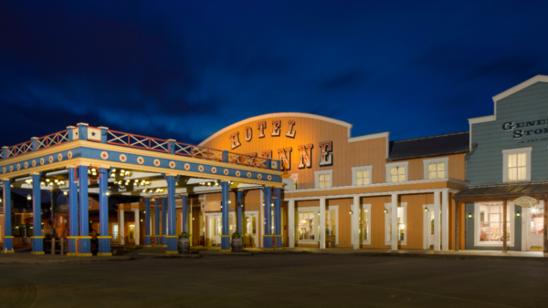 Vue nocturne d'une place de ville à thème western, avec un bâtiment étiqueté « Hôtel Cheyenne Disney » et un « Magasin général », avec des guirlandes lumineuses illuminant la zone, promettant un séjour mémorable.