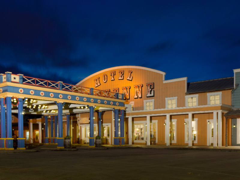 Vue nocturne d'une place de ville à thème western, avec un bâtiment étiqueté « Hôtel Cheyenne Disney » et un « Magasin général », avec des guirlandes lumineuses illuminant la zone, promettant un séjour mémorable.