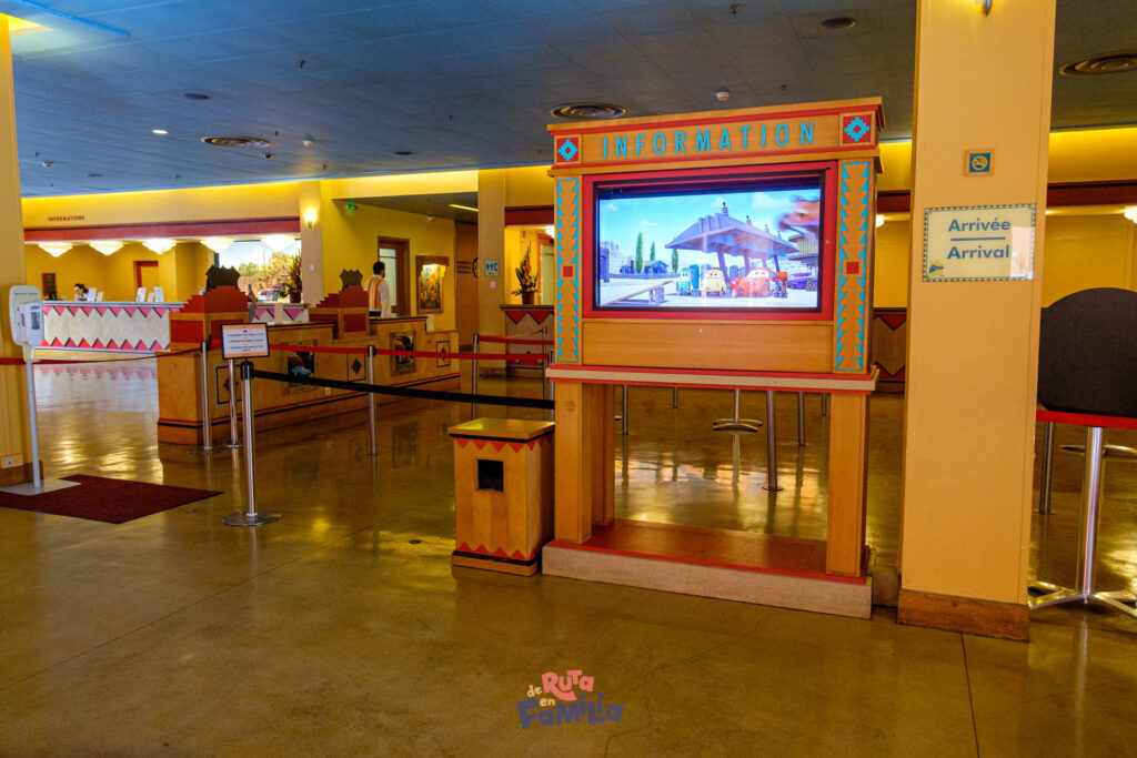 Un stand d'information avec écran numérique dans un espace intérieur lumineux, avec une zone de file d'attente et une signalisation d'arrivée. Différents comptoirs et décors sont visibles en arrière-plan, rappelant l'atmosphère animée du Disney Hotel Santa Fe.