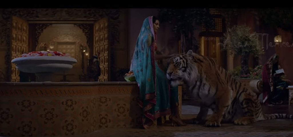 Une femme en tenue traditionnelle se tient à côté d'un tigre dans une pièce richement décorée avec des meubles ornés et un bol de fruits, évoquant l'ambiance enchanteresse des nuits d'arabie.