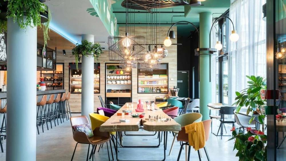 Un café moderne avec des chaises colorées, une table en bois centrale, un éclairage suspendu et un coin bar en arrière-plan. La lumière naturelle et la lumière naturelle rehaussent le cadre, lui donnant une ambiance chaleureuse de restaurant Goma en plein Val d'Europe.