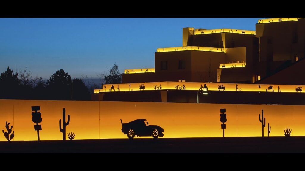 Silhouette d'un mur orné de décorations sur le thème du désert et d'une silhouette de voiture, sur fond d'un bâtiment à plusieurs niveaux illuminé au crépuscule. Bienvenue à l'hôtel Santa Fe, démarrez votre moteur pour un séjour inoubliable !