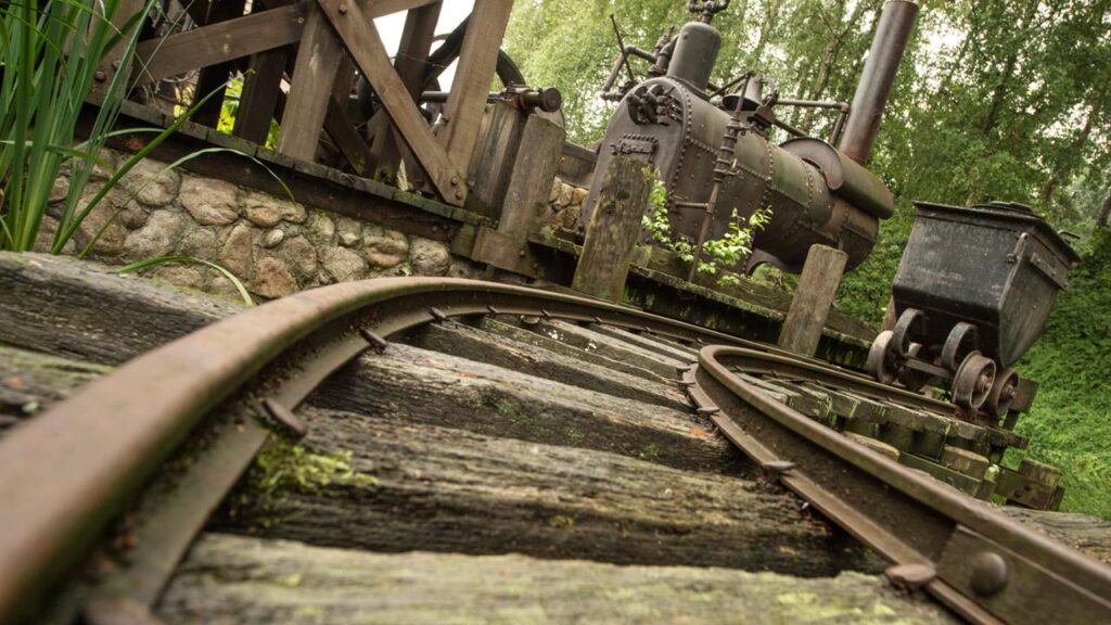 Vue rapprochée de voies ferrées rouillées et courbes rappelant Big Thunder Mountain menant à une vieille machine industrielle et à un petit chariot, entourés de verdure.