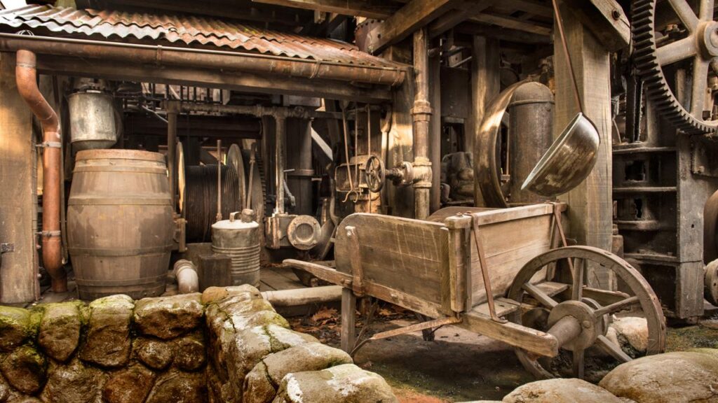 Un atelier rustique à l'ancienne avec des tonneaux en bois, des tuyaux en métal, des engrenages et une brouette en bois au milieu de fondations en pierre et de machines, rappelant le charme robuste de Big Thunder Mountain.