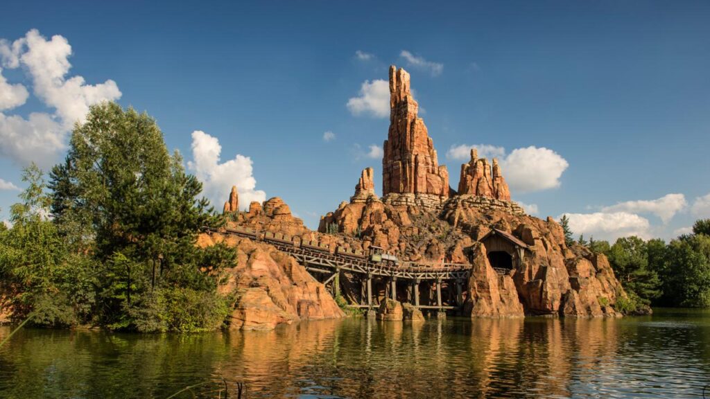 Des montagnes russes serpentent à travers le terrain rocheux de Big Thunder Mountain, entourées d'arbres et d'un plan d'eau scintillant sous un ciel bleu parsemé de nuages.