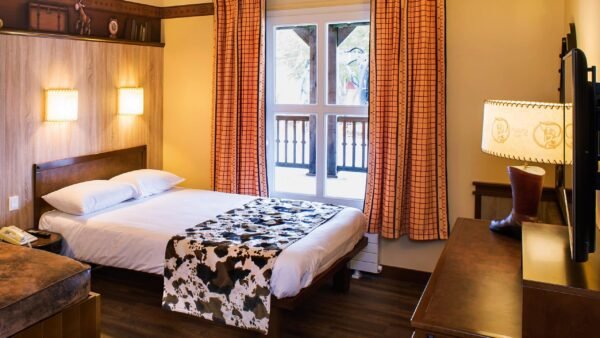 Une chambre confortable et bien éclairée avec un lit double, une table de chevet et une fenêtre avec des rideaux tirés. La chambre dispose d'un éclairage chaleureux et d'une décoration rustique inspirée de la culture Cheyenne, notamment un chemin de lit à motif de vache.
