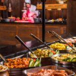 Une table de buffet est dressée avec divers plats, notamment des légumes, des fruits de mer et de la viande. Un chef découpe de la viande en arrière-plan. Découvrez ce festin gastronomique dans le meilleur lieu de restauration de Disney Village en 2024.