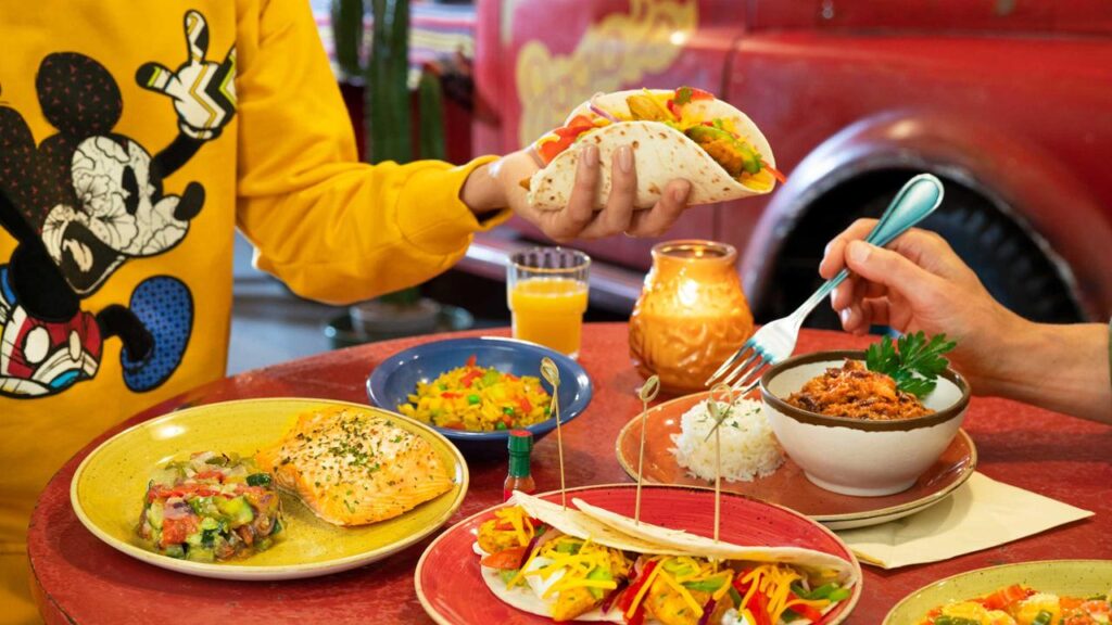 Une table remplie de plats variés, dont des tacos, des enchiladas, du riz aux haricots, du saumon et du pico de gallo. Une personne portant une chemise jaune tient un taco tandis qu'une autre personne utilise une fourchette. La scène animée ressemble à une fête tout droit sortie de l'Hôtel Santa Fe au bord du Rio Grande.