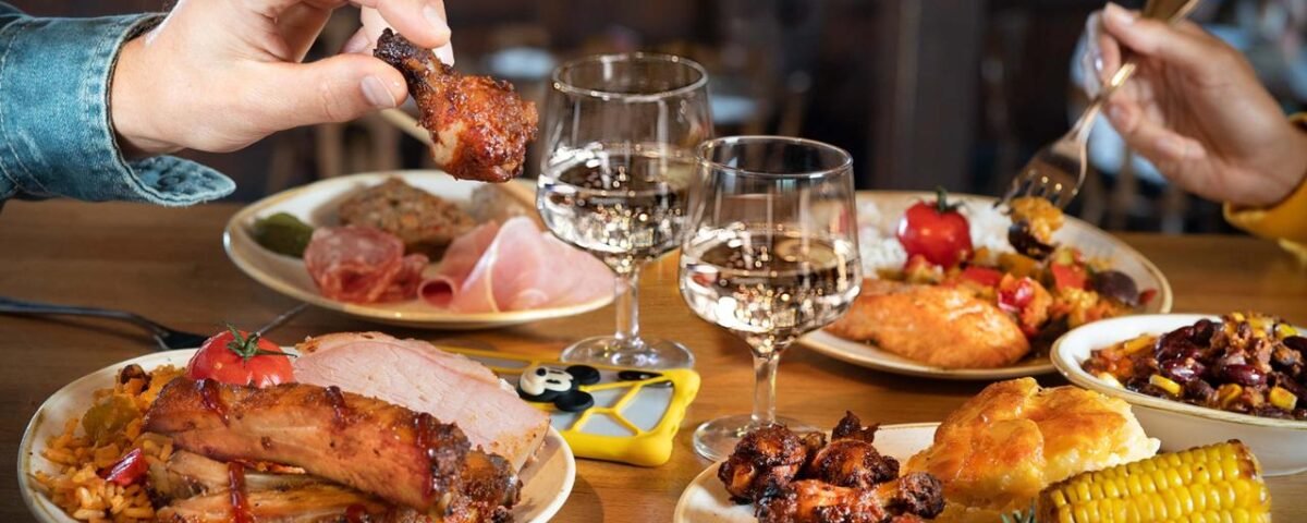 Deux personnes dégustent un repas composé de plats variés, notamment du poulet grillé, des épis de maïs, des côtes levées et des légumes, dans un restaurant chaleureux de Disney Village. Deux verres de vin blanc sont sur la table. La scène promet des expériences de restauration délicieuses qui devraient s'élever en 2024.