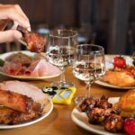 Deux personnes dégustent un repas composé de plats variés, notamment du poulet grillé, des épis de maïs, des côtes levées et des légumes, dans un restaurant chaleureux de Disney Village. Deux verres de vin blanc sont sur la table. La scène promet des expériences de restauration délicieuses qui devraient s'élever en 2024.