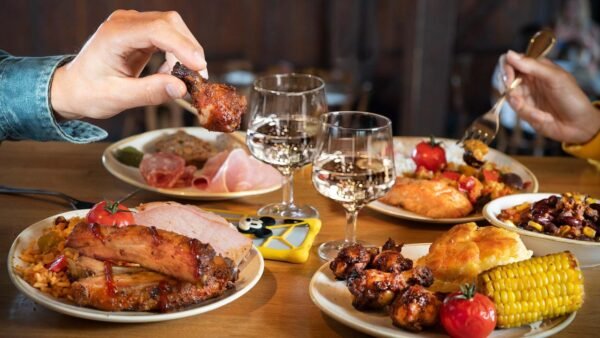 Deux personnes dégustent un repas composé de plats variés, notamment du poulet grillé, des épis de maïs, des côtes levées et des légumes, dans un restaurant chaleureux de Disney Village. Deux verres de vin blanc sont sur la table. La scène promet des expériences de restauration délicieuses qui devraient s'élever en 2024.