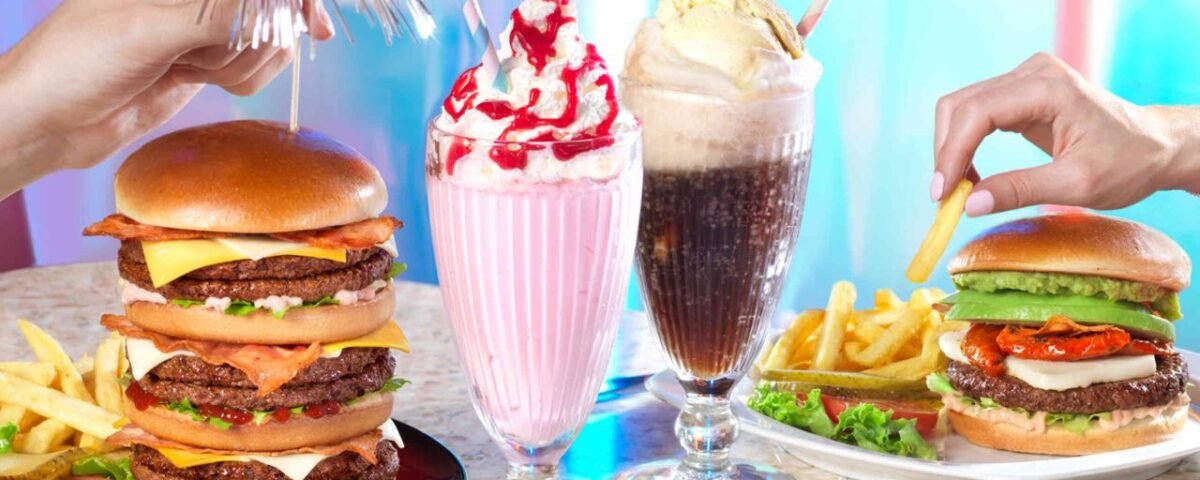 Deux mains tenant un double cheeseburger et un milkshake, une autre main tenant une frite près d'un burger végétarien avec des frites. Deux milkshakes garnis de crème fouettée et de cerises sont placés entre eux au nouveau point de restauration de Disney Village 2024.