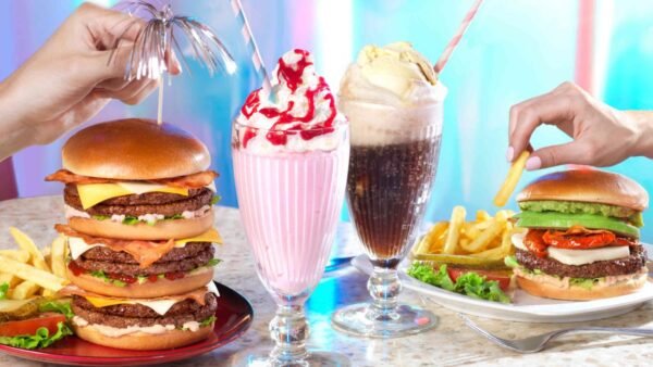 Deux mains tenant un double cheeseburger et un milkshake, une autre main tenant une frite près d'un burger végétarien avec des frites. Deux milkshakes garnis de crème fouettée et de cerises sont placés entre eux au nouveau point de restauration de Disney Village 2024.