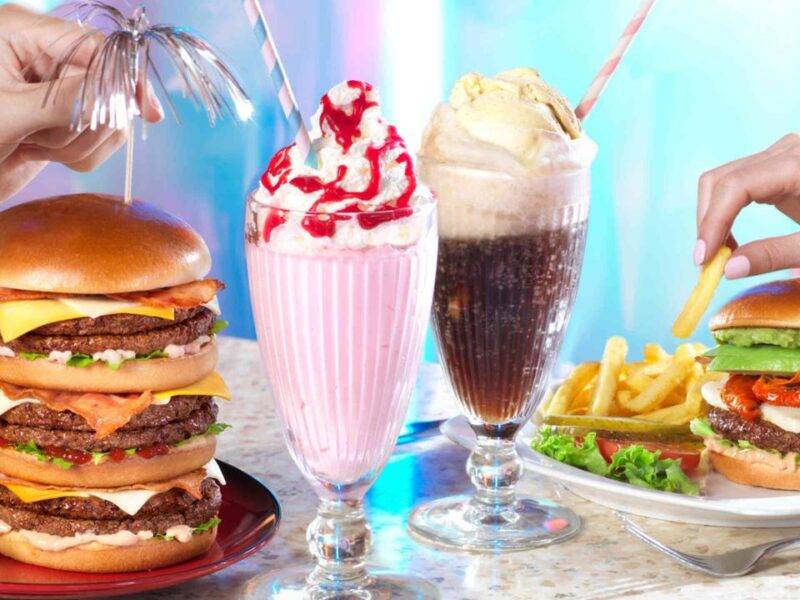 Deux mains tenant un double cheeseburger et un milkshake, une autre main tenant une frite près d'un burger végétarien avec des frites. Deux milkshakes garnis de crème fouettée et de cerises sont placés entre eux au nouveau point de restauration de Disney Village 2024.