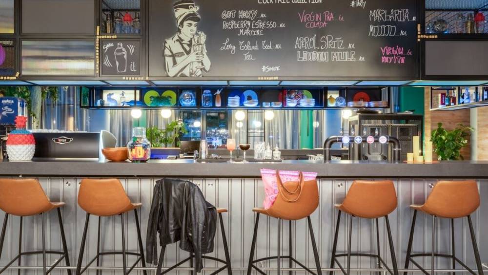 Un bar avec un comptoir en tôle ondulée, quatre tabourets en cuir marron, une veste drapée sur l'un d'eux et diverses boissons et décorations sur le comptoir. Un menu à la craie est visible au-dessus du bar, promettant une expérience culinaire inoubliable.