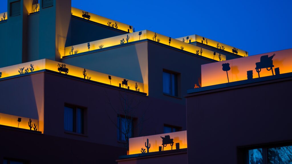 Un bâtiment moderne avec des terrasses décorées de sculptures en silhouette est illuminé par des lumières jaunes chaudes au crépuscule, rappelant l'élégance stylisée du Newport Bay Club.