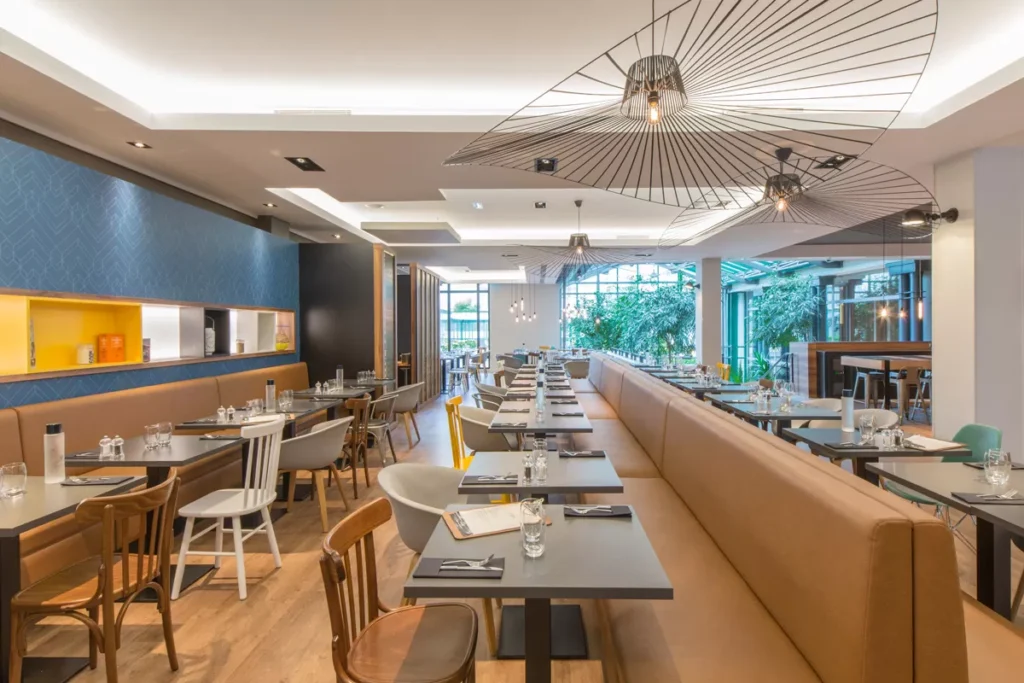 Un intérieur de restaurant moderne avec des tables et des chaises soigneusement disposées. L'espace est bien éclairé par des plafonniers géométriques uniques et de grandes fenêtres laissent entrer la lumière naturelle, rappelant l'ambiance chic de Val d'Europe.