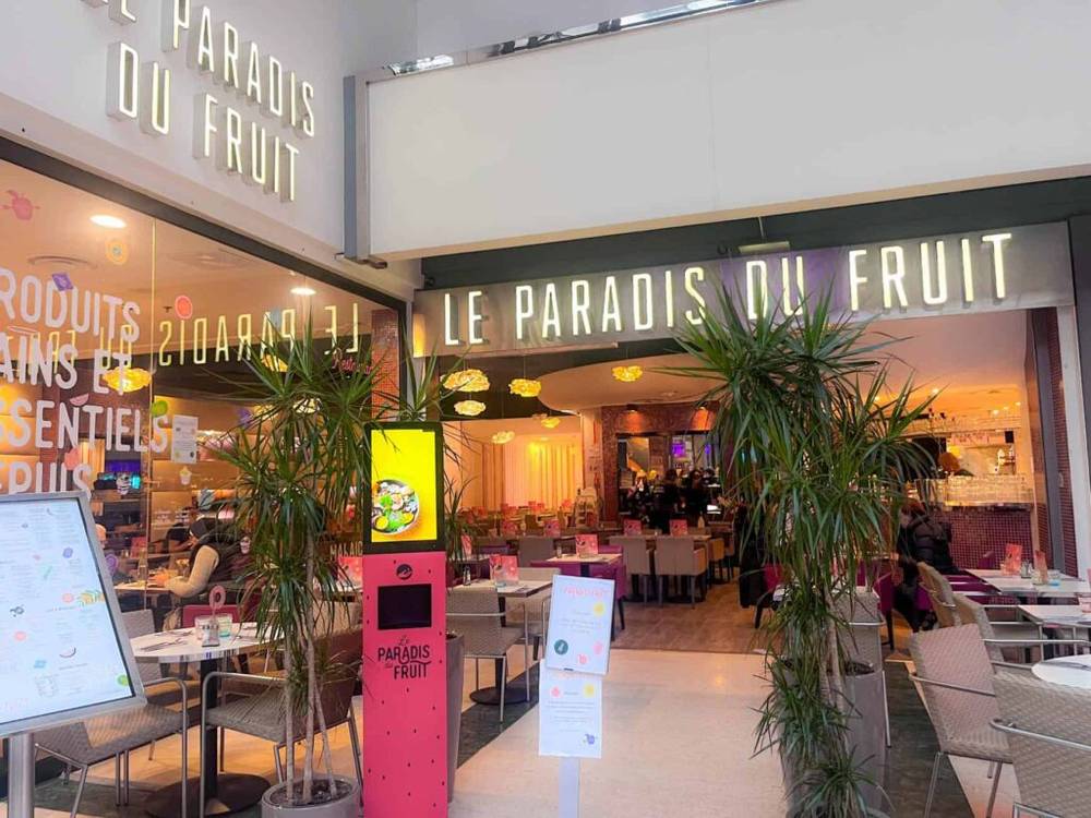 Vue extérieure du restaurant "Le Paradis du Fruit" à Val d'Europe avec ses murs en verre, ses plantes en pot et ses menus affichés. L'intérieur est très éclairé et décoré de meubles modernes et de luminaires suspendus.