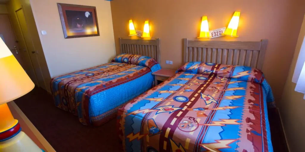 Une chambre d'hôtel avec deux lits simples, chacun avec une literie aux motifs lumineux. La chambre présente un thème coloré rappelant l'Hôtel Santa Fe, avec des lumières murales et un tableau encadré au-dessus des lits. Démarrez votre moteur et profitez de votre séjour !