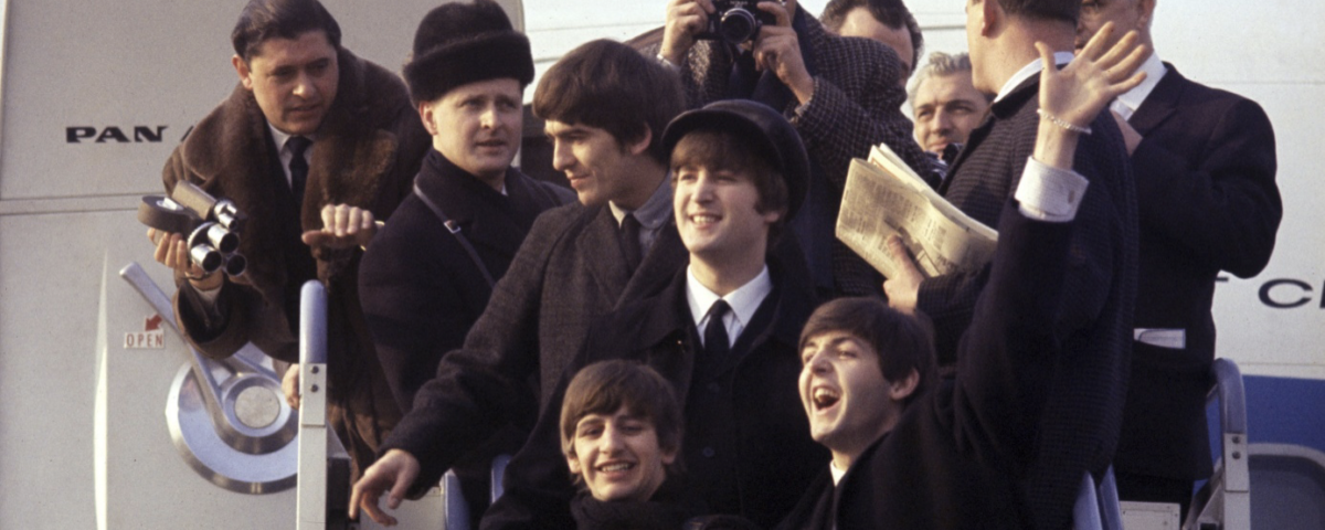 Un groupe de personnes pose et salue en haut des escaliers d'un avion, rappelant l'arrivée des Beatles à l'aéroport, avec des photographes et des spectateurs capturant le moment comme une scène d'une aventure Disney.