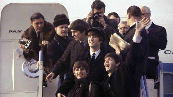 Un groupe de personnes pose et salue en haut des escaliers d'un avion, rappelant l'arrivée des Beatles à l'aéroport, avec des photographes et des spectateurs capturant le moment comme une scène d'une aventure Disney.