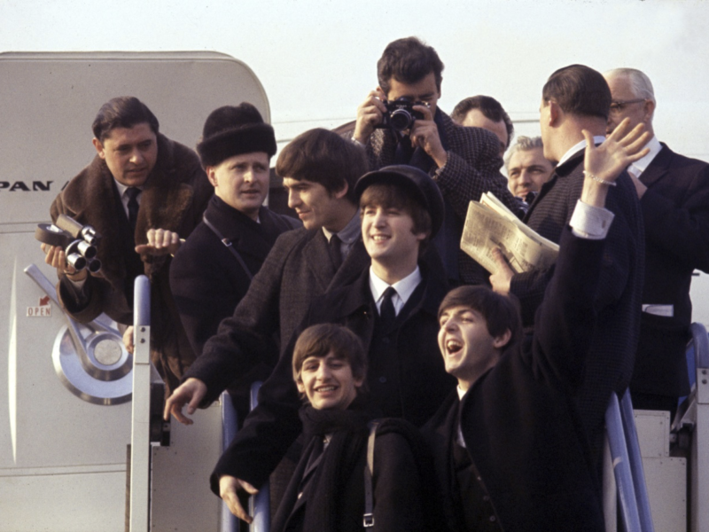 Un groupe de personnes pose et salue en haut des escaliers d'un avion, rappelant l'arrivée des Beatles à l'aéroport, avec des photographes et des spectateurs capturant le moment comme une scène d'une aventure Disney.