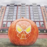 Grande citrouille avec un motif de masque d'araignée stylisé sur le devant, placée devant un bâtiment moderne à plusieurs étages, capturant l'esprit enchanteur du Mode Halloween que vous trouverez à Disneyland.