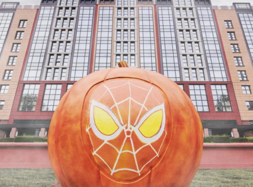 Grande citrouille avec un motif de masque d'araignée stylisé sur le devant, placée devant un bâtiment moderne à plusieurs étages, capturant l'esprit enchanteur du Mode Halloween que vous trouverez à Disneyland.