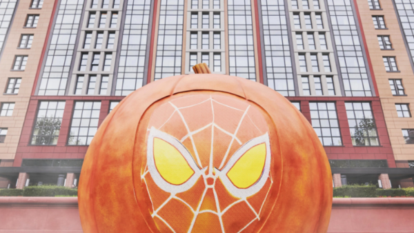 Grande citrouille avec un motif de masque d'araignée stylisé sur le devant, placée devant un bâtiment moderne à plusieurs étages, capturant l'esprit enchanteur du Mode Halloween que vous trouverez à Disneyland.
