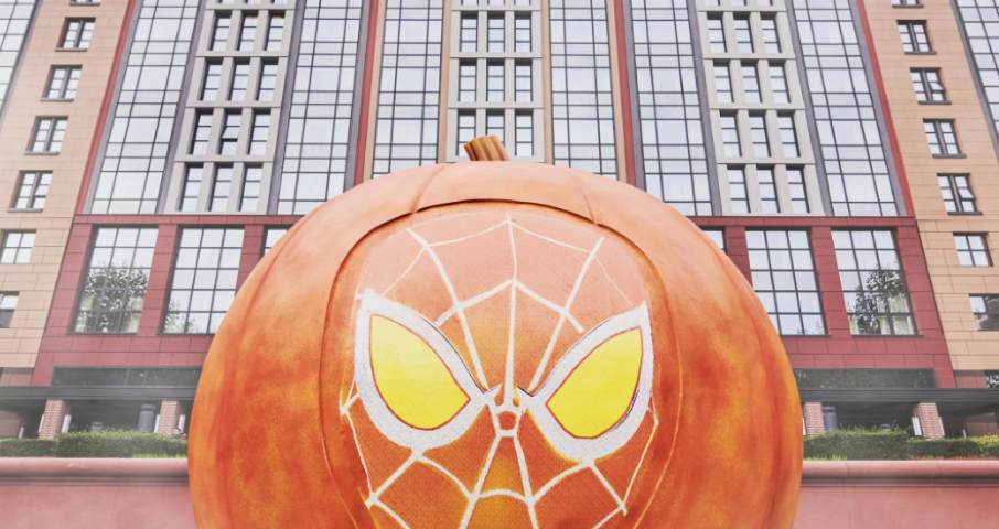 Grande citrouille avec un motif de masque d'araignée stylisé sur le devant, placée devant un bâtiment moderne à plusieurs étages, capturant l'esprit enchanteur du Mode Halloween que vous trouverez à Disneyland.