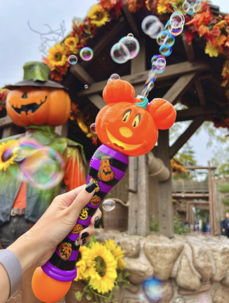 Une main tient une baguette à bulles sur le thème d'Halloween avec un motif de citrouille fantaisiste, rappelant un objet de collection à échanger des broches. Les bulles flottent autour, se mêlant à un épouvantail et à des décorations d'automne en arrière-plan.